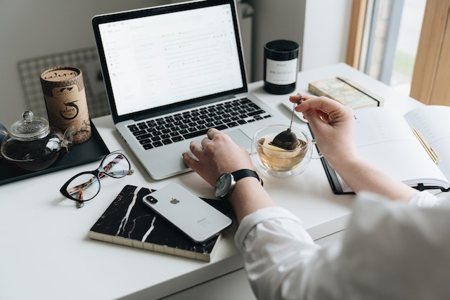 Person's hands at a desk that is set up with a laptop, notebook, and cup of tea