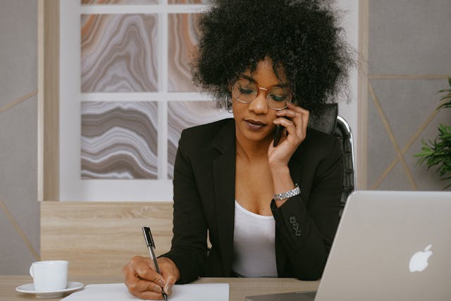 woman in a black blazer talking on a phone and writing notes